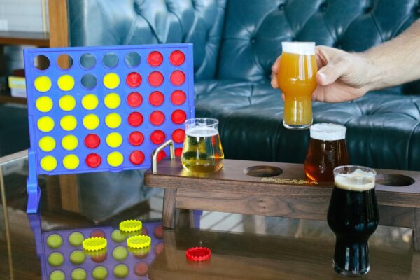 A Connect Four game with red and yellow pieces sits on a table next to a wooden beer flight holder with three different beers. A person’s hand is picking up a glass of orange-colored beer. Bottle caps are scattered on the table.