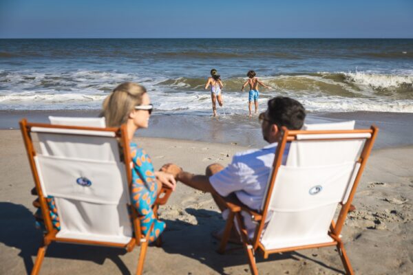 A man and woman sit in beach chairs facing the ocean, holding hands, while two children play at the edge of the water under a clear blue sky.