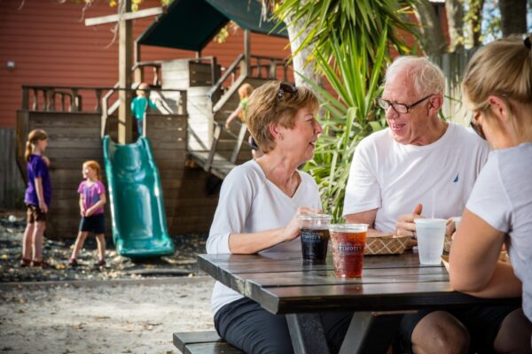 Two older adults sit at an outdoor picnic table, smiling and talking, with drinks and food in front of them. A playground with children playing is visible in the background on a sunny day.