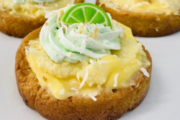 A close-up of a cookie topped with creamy yellow filling, toasted coconut flakes, green whipped cream, and a small slice of lime, creating a key lime pie-inspired dessert.