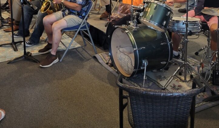 A jazz band performs indoors, with musicians playing saxophones, brass, and drums. The group includes men and women of various ages. A sign in front reads, Nassau Community Jazz Band with a trumpet graphic.