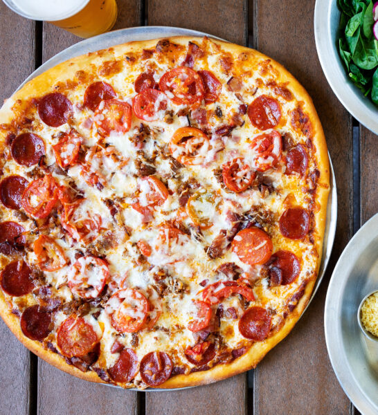 A large pizza topped with pepperoni, sliced tomatoes, cheese, and bacon on a wooden table at Natural Slice in Omni Amelia Island Resort, beside a plate of salad, a drink, and a small dish of grated cheese.