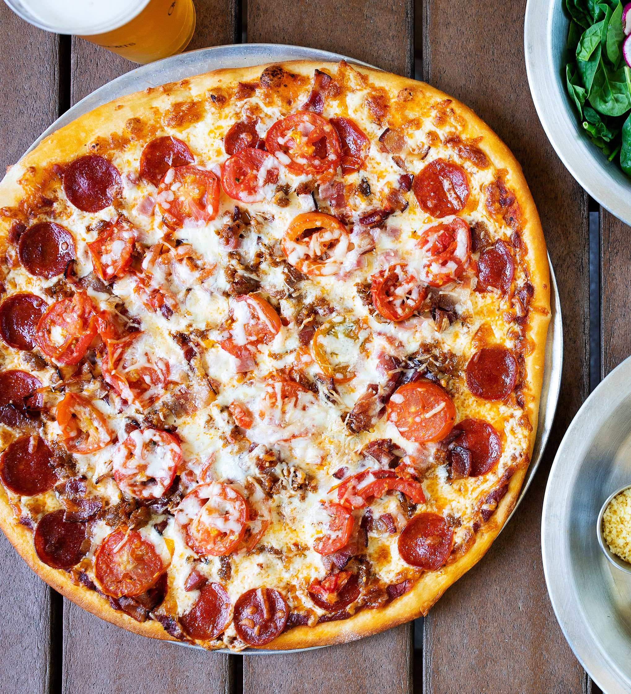A large pizza topped with pepperoni, sliced tomatoes, cheese, and bacon on a wooden table at Natural Slice in Omni Amelia Island Resort, beside a plate of salad, a drink, and a small dish of grated cheese.