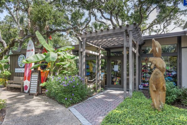 The image shows the entrance to a nature gift shop surrounded by lush greenery, with a pergola, carved wooden sculpture, surfboard, and colorful displays visible through the glass doors.