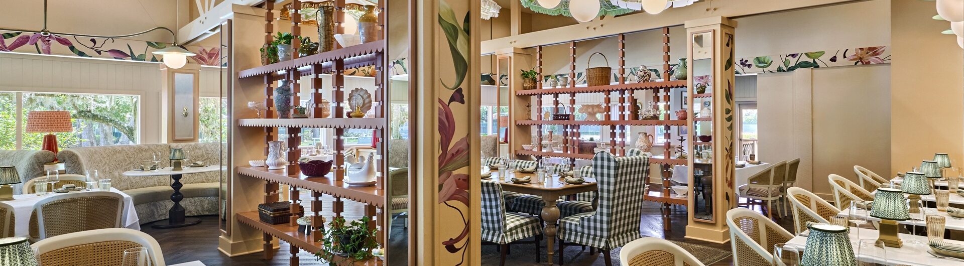 A bright, elegant restaurant with floral decor, wooden floors, white marble tables, wicker chairs, and green glass lamps. A wooden divider with plants separates two dining areas, each with cozy seating and colorful painted walls.