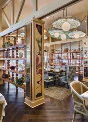 A bright, elegant restaurant with floral decor, wooden floors, white marble tables, wicker chairs, and green glass lamps. A wooden divider with plants separates two dining areas, each with cozy seating and colorful painted walls.