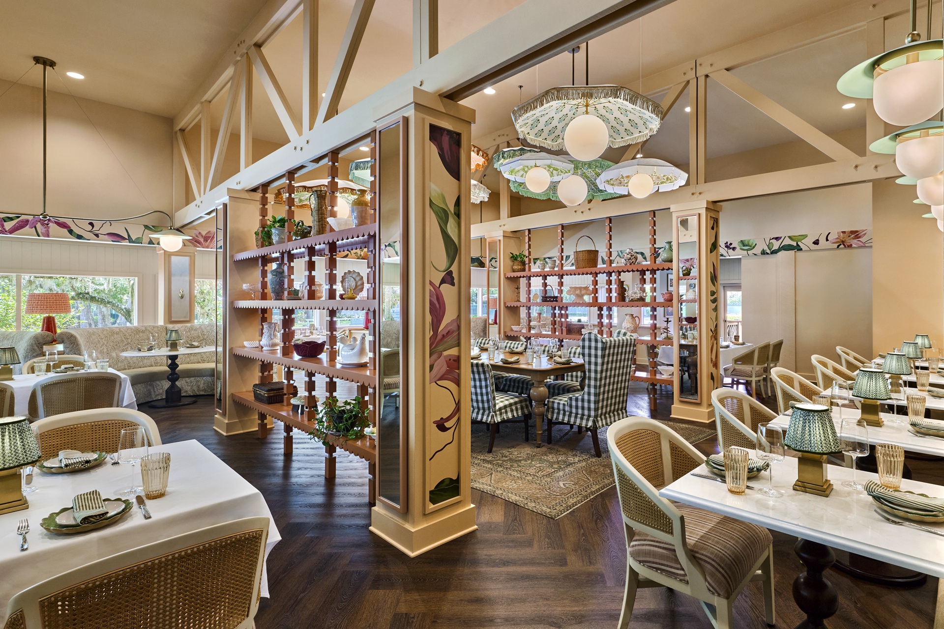 A bright, elegant restaurant with floral decor, wooden floors, white marble tables, wicker chairs, and green glass lamps. A wooden divider with plants separates two dining areas, each with cozy seating and colorful painted walls.