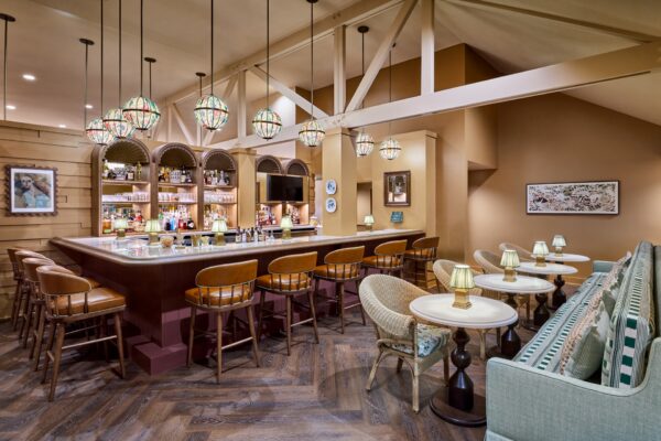 Cozy bar with wooden stools, cushioned seating, hanging stained glass lamps, and decorative art on beige walls. Bottled drinks line the bar shelves under arched niches.