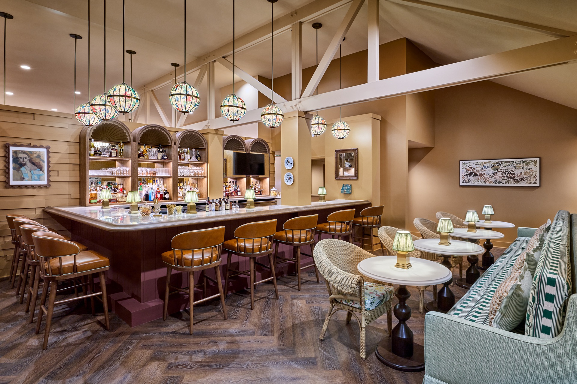 Cozy bar with wooden stools, cushioned seating, hanging stained glass lamps, and decorative art on beige walls. Bottled drinks line the bar shelves under arched niches.