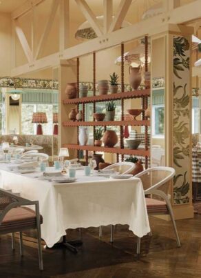 A bright, elegant restaurant interior with white tablecloths, checkered cushioned chairs, wooden floors, decorative shelving with plants and vases, and large windows letting in natural light.