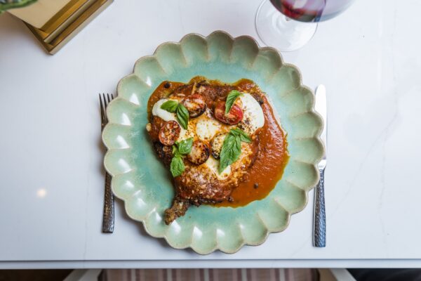 A scalloped plate holds a breaded meat cutlet topped with tomato sauce, mozzarella, cherry tomatoes, and basil. A fork, knife, and glass of red wine are set beside it on a white table.