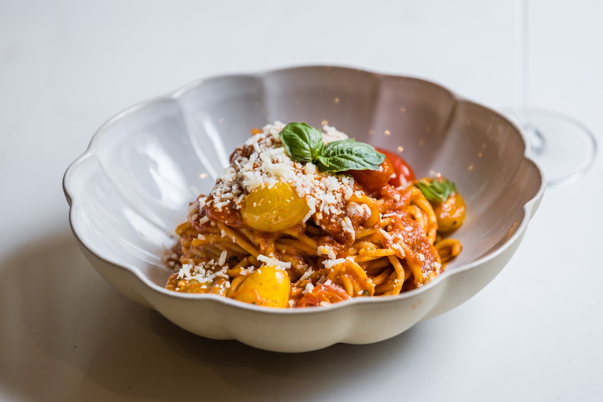 A bowl of spaghetti topped with tomato sauce, yellow and red cherry tomatoes, grated cheese, and a fresh basil leaf, served in a white scalloped bowl.