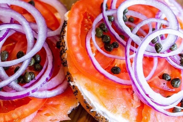 A close-up of a bagel topped with cream cheese, slices of smoked salmon, tomato, red onion rings, and capers.