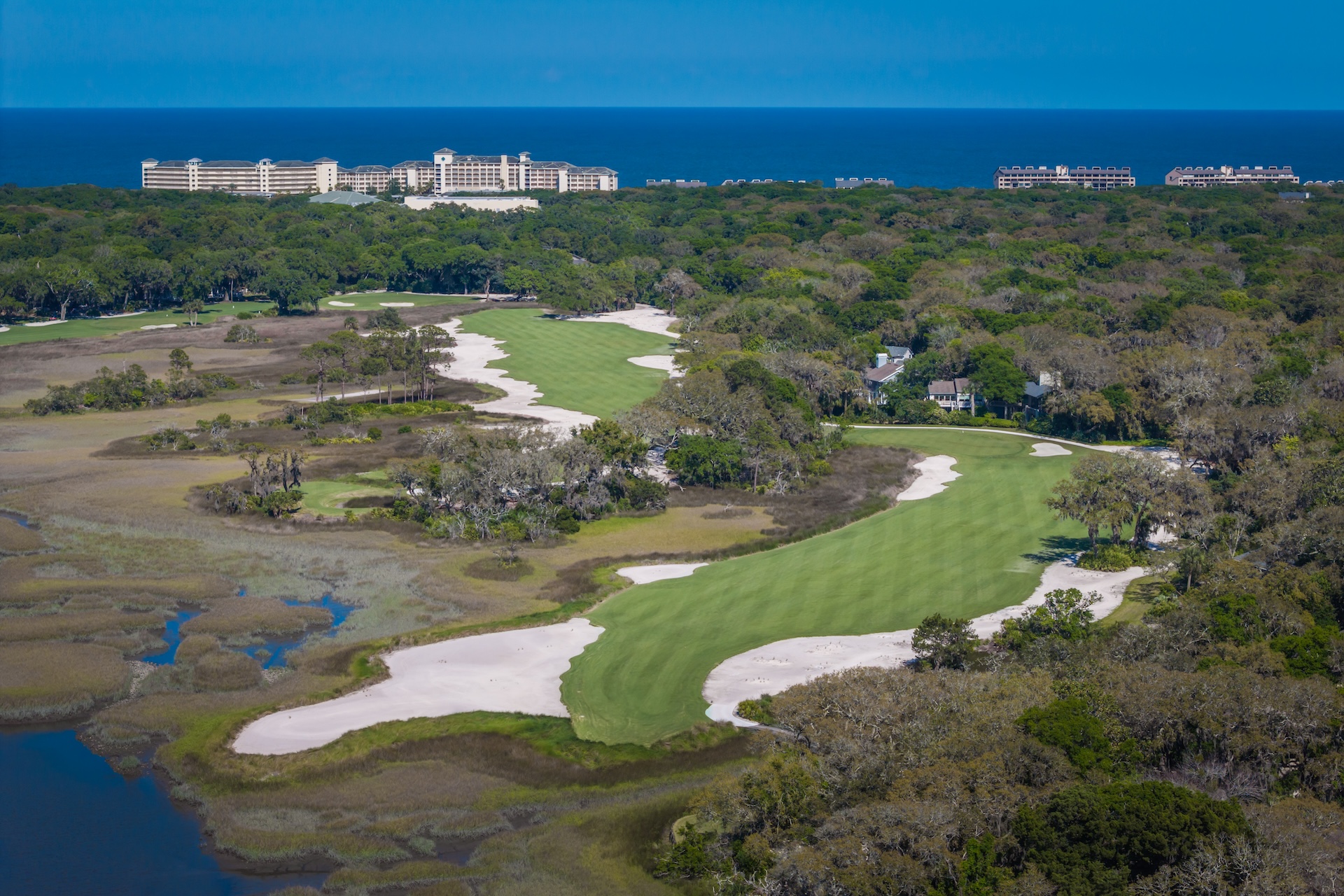 Aerial view of a coastal golf course featuring winding green fairways and sand bunkers, surrounded by forest and marshland, with large resort buildings near the ocean in the distance.
