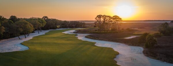 A scenic golf course at sunset with a green fairway bordered by white sand bunkers, trees on the left, and open marshland on the right under a glowing orange sky.