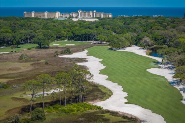 Aerial view of a long golf course fairway winding through dense forest and sand bunkers, with a large resort hotel and the ocean in the distance.