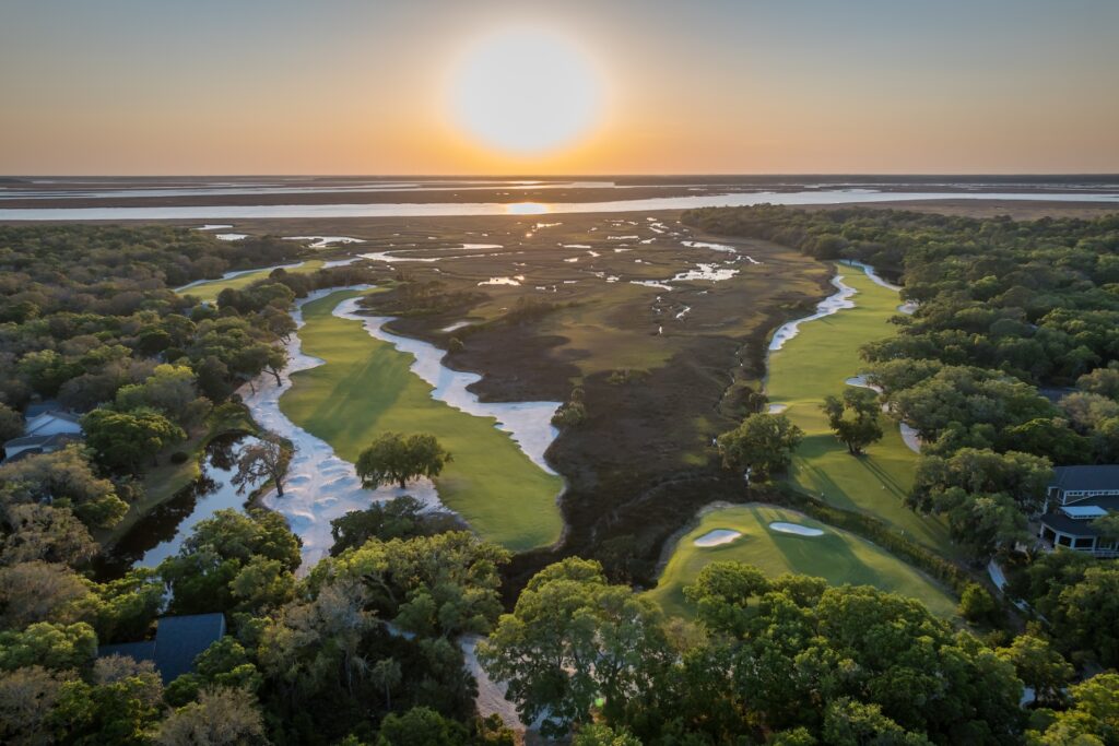 Aerial view of a lush golf course at sunset, with green fairways bordered by trees and sand traps, winding alongside a marshy waterway. The sun sets over the distant horizon, casting a golden glow across the landscape.