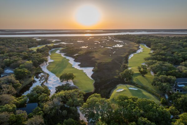 Aerial view of a lush golf course at sunset, with green fairways bordered by trees and sand traps, winding alongside a marshy waterway. The sun sets over the distant horizon, casting a golden glow across the landscape.