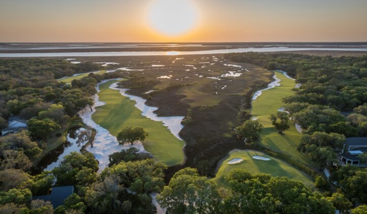 Aerial view of a lush golf course at sunset, with green fairways bordered by trees and sand traps, winding alongside a marshy waterway. The sun sets over the distant horizon, casting a golden glow across the landscape.