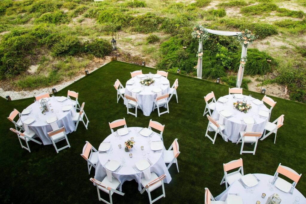 Six round tables with white tablecloths and floral centerpieces are arranged on a green lawn, each surrounded by white chairs. An arch decorated with flowers stands at the edge, with greenery in the background.