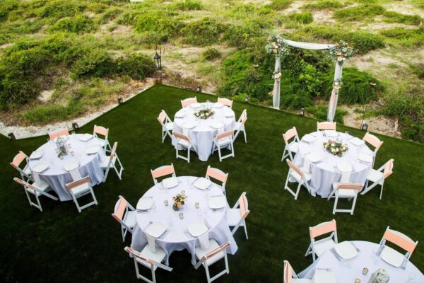 Six round tables with white tablecloths and floral centerpieces are arranged on a green lawn, each surrounded by white chairs. An arch decorated with flowers stands at the edge, with greenery in the background.