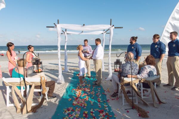 A couple stands under a white canopy on a beach, holding hands and facing each other while a person officiates. Guests are seated on either side, with colorful petals scattered along a turquoise aisle.