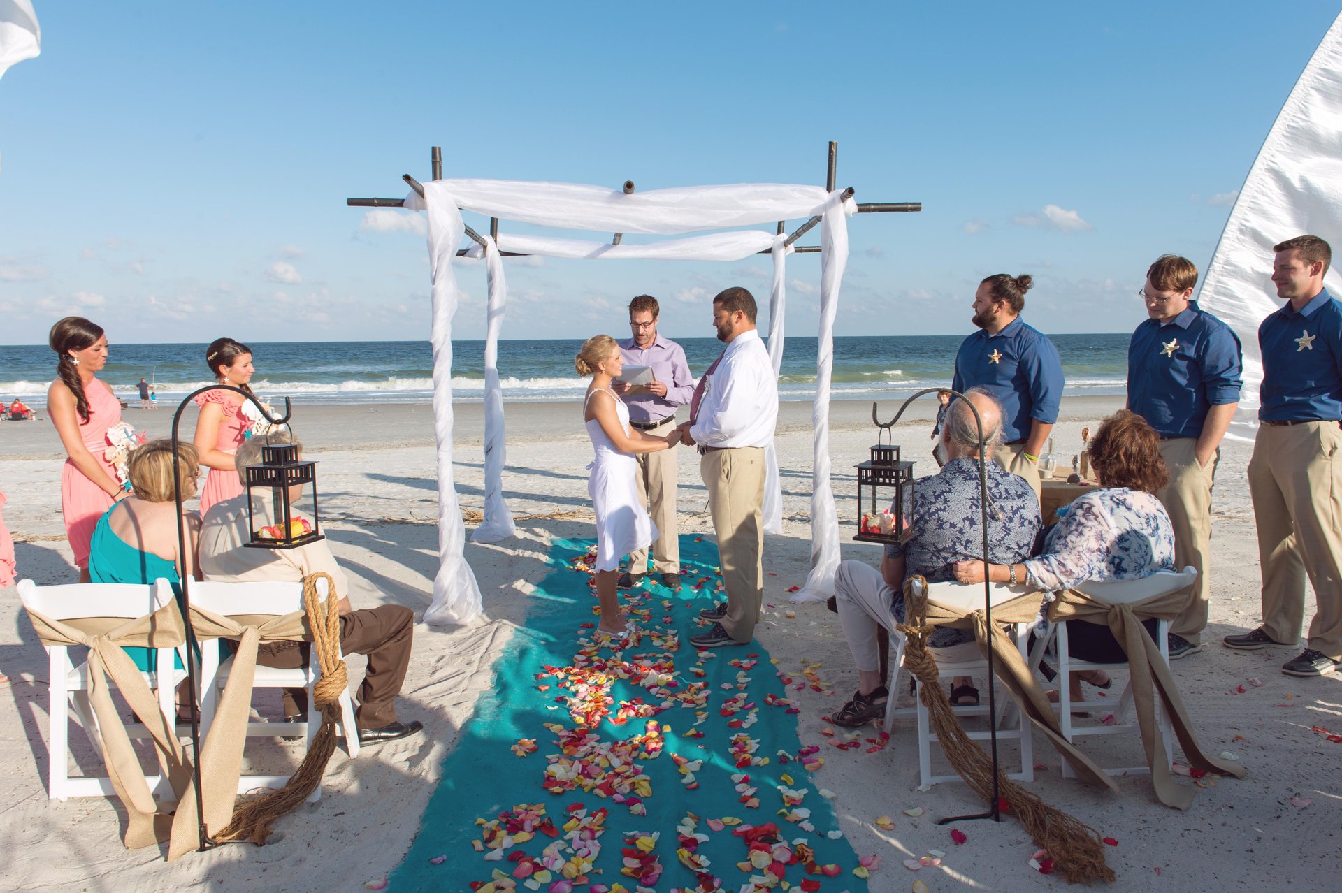 A couple stands under a white canopy on a beach, holding hands and facing each other while a person officiates. Guests are seated on either side, with colorful petals scattered along a turquoise aisle.