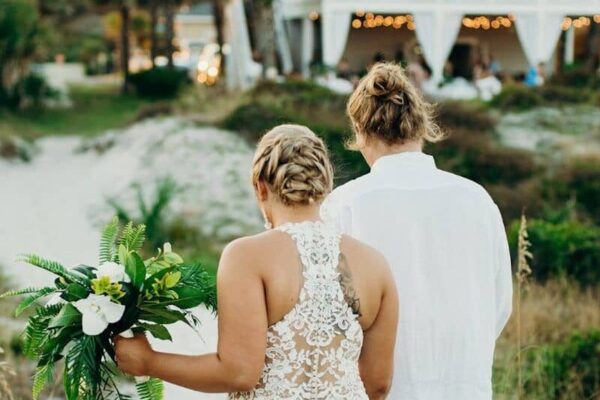 A bride holding a bouquet stands next to a person in a white shirt; both are seen from behind, outdoors, with greenery, sand, and a decorated venue in the background.
