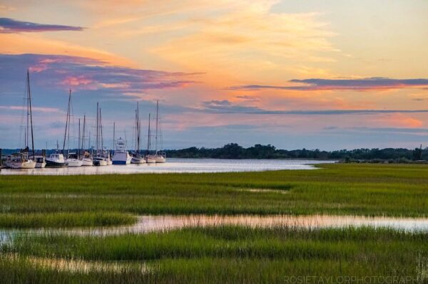 Sailboats docked along a calm river at sunset, with colorful clouds in the sky and green marsh grasses in the foreground.