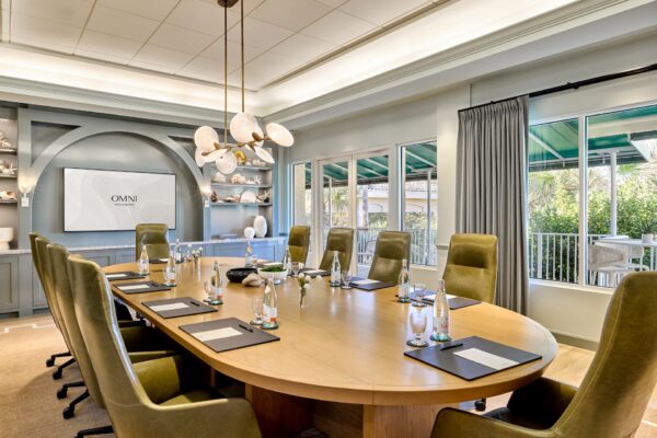 Modern conference room with a long wooden table, green chairs, bottled water, notepads, and pens. Large windows let in natural light. A screen displays Omni. Shelves with decor are on the left wall.