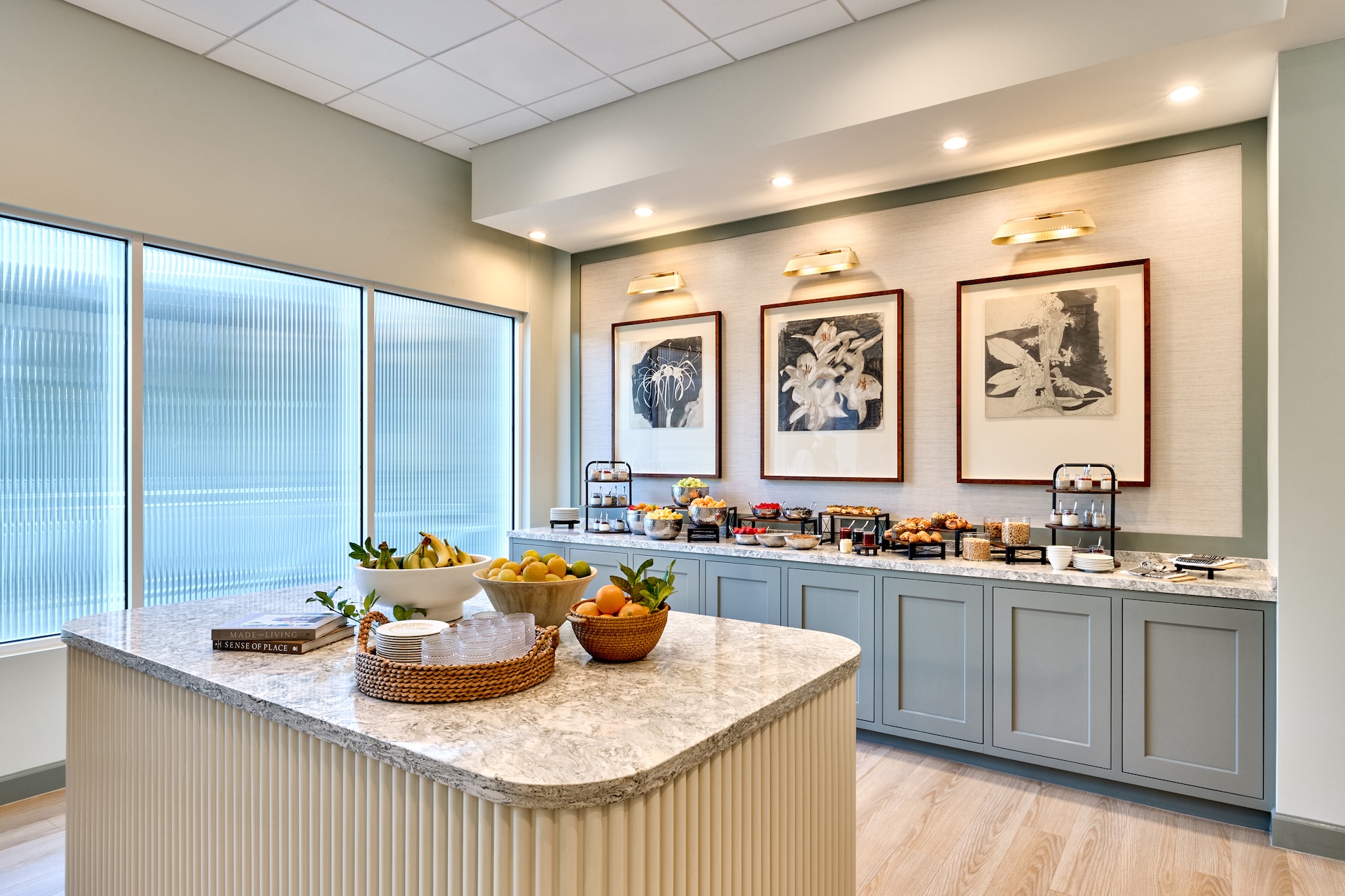 A bright, modern kitchen features a marble island with fruit bowls, cabinets lined with breakfast items, coffee makers, and framed art on the wall above. Large windows provide natural light.
