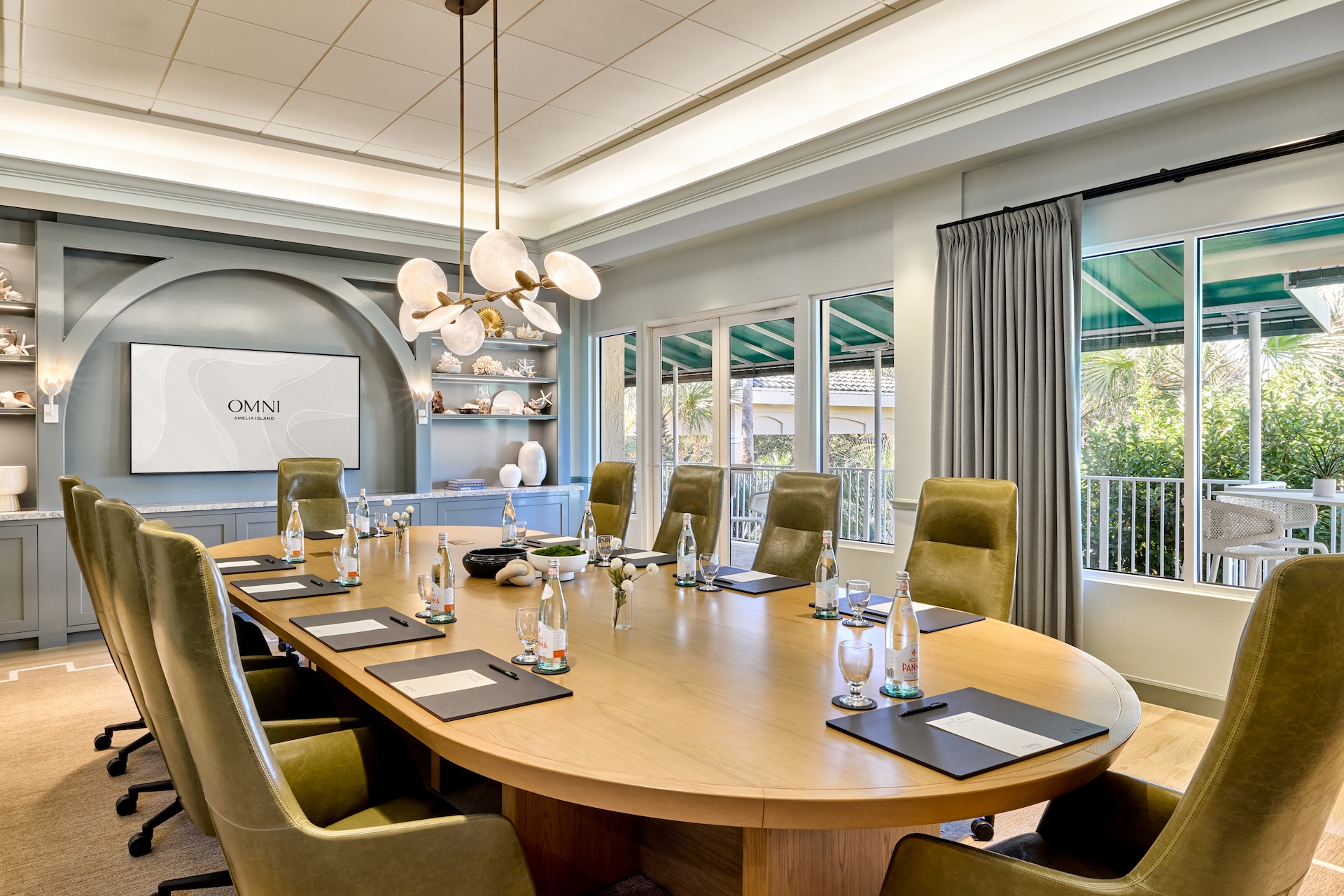 Modern conference room with a long wooden table, green chairs, bottled water, notepads, and pens. Large windows let in natural light. A screen displays Omni. Shelves with decor are on the left wall.