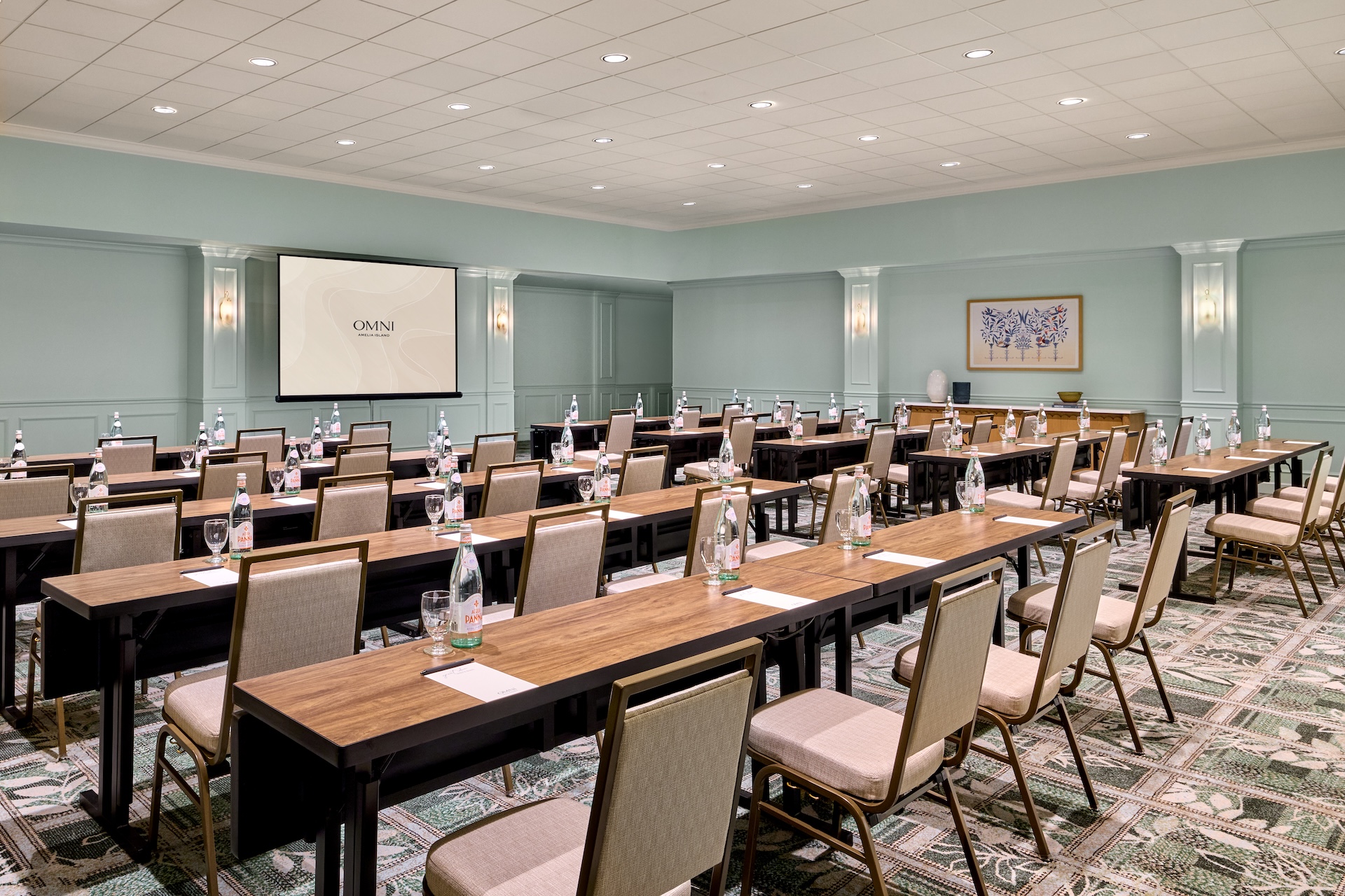Conference room with rows of tables and chairs facing a projector screen. Each table has water bottles, glasses, and notepads. The room has pale walls, patterned carpet, and framed artwork on the back wall.