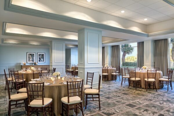 Elegant banquet room with round tables covered in brown cloths, set with glassware, white floral centerpieces, and wooden chairs. Large windows let in natural light and offer a view of palm trees outside.