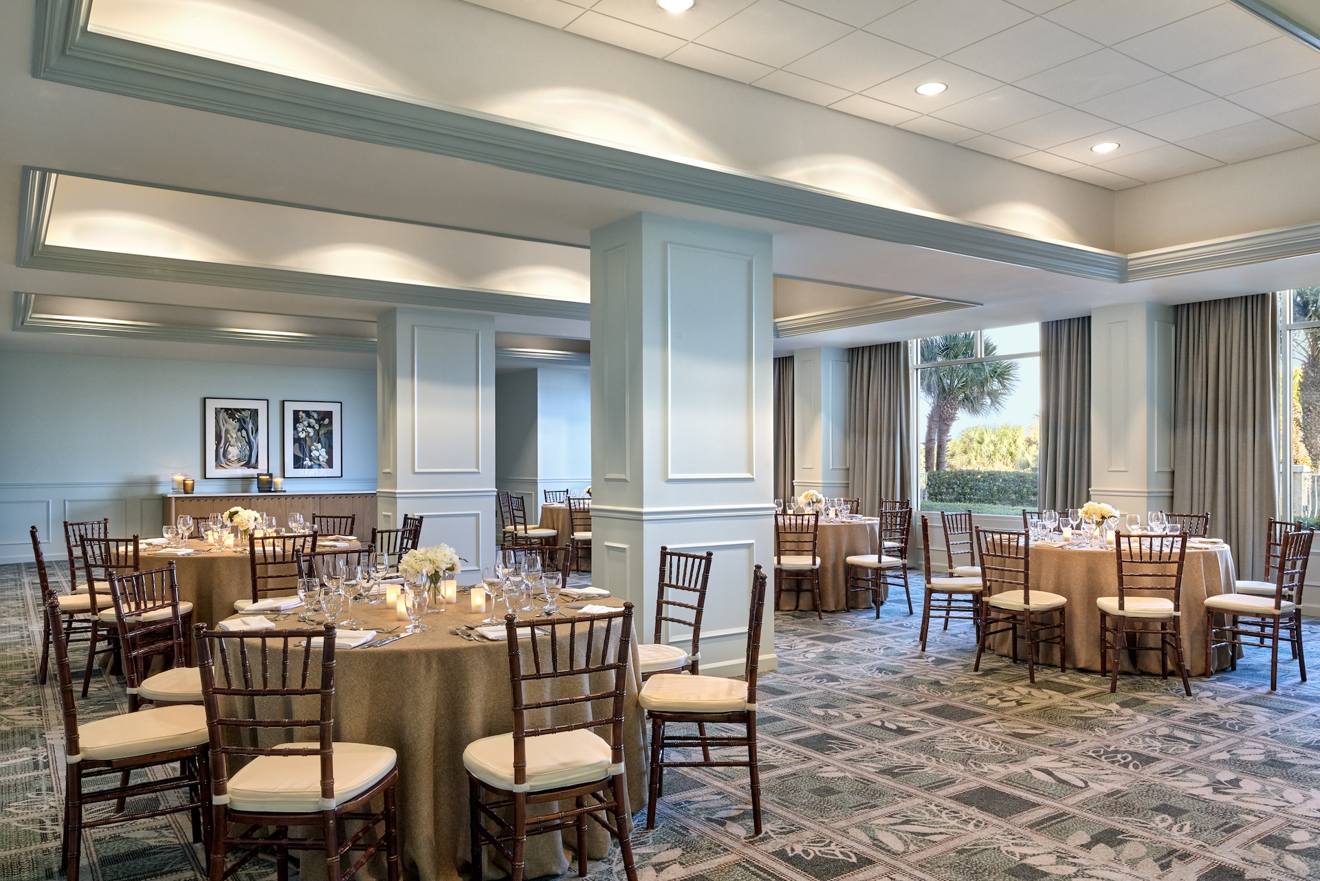 Elegant banquet room with round tables covered in brown cloths, set with glassware, white floral centerpieces, and wooden chairs. Large windows let in natural light and offer a view of palm trees outside.