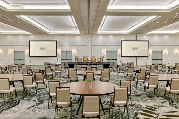 Large, modern conference room with round tables and beige chairs facing a stage. Two large screens display OMNI. The space is well-lit, with patterned carpet and a high ceiling.