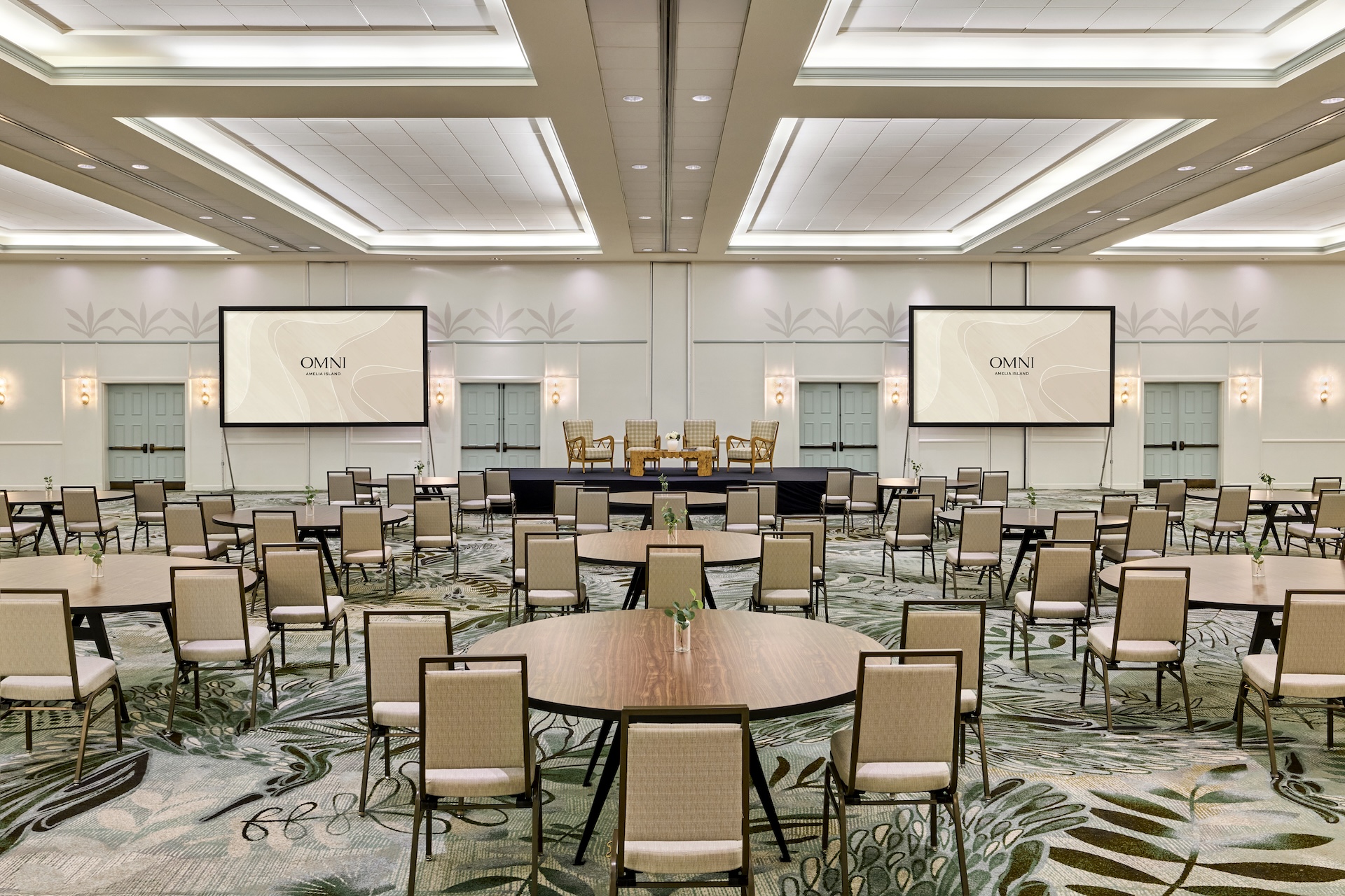 Large, modern conference room with round tables and beige chairs facing a stage. Two large screens display OMNI. The space is well-lit, with patterned carpet and a high ceiling.