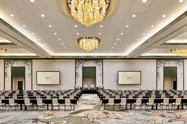 Spacious, elegant conference room with rows of chairs facing a stage and four screens, illuminated by large chandeliers, floral carpet, and decorative wall panels.