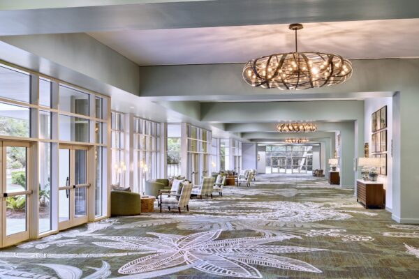 Spacious hotel lobby with large windows, patterned carpet, modern chandeliers, cozy seating areas, green armchairs, and natural light streaming in.