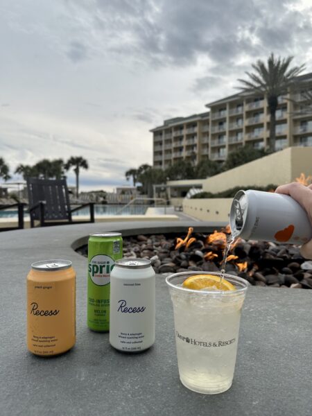 A hand pours a drink into a cup with orange slices by a fire pit at a resort poolside. Three canned beverages—Recess Peach Ginger, Recess Coconut Lime, and Melon Mint Sprite—are on the ledge.