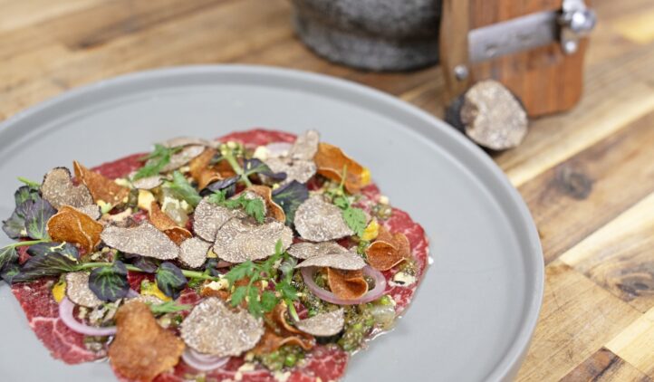 A gourmet dish of thinly sliced raw beef topped with shaved truffles, herbs, pickled onions, and crispy vegetable chips, served on a gray plate on a wooden table. A truffle and slicer are in the background.