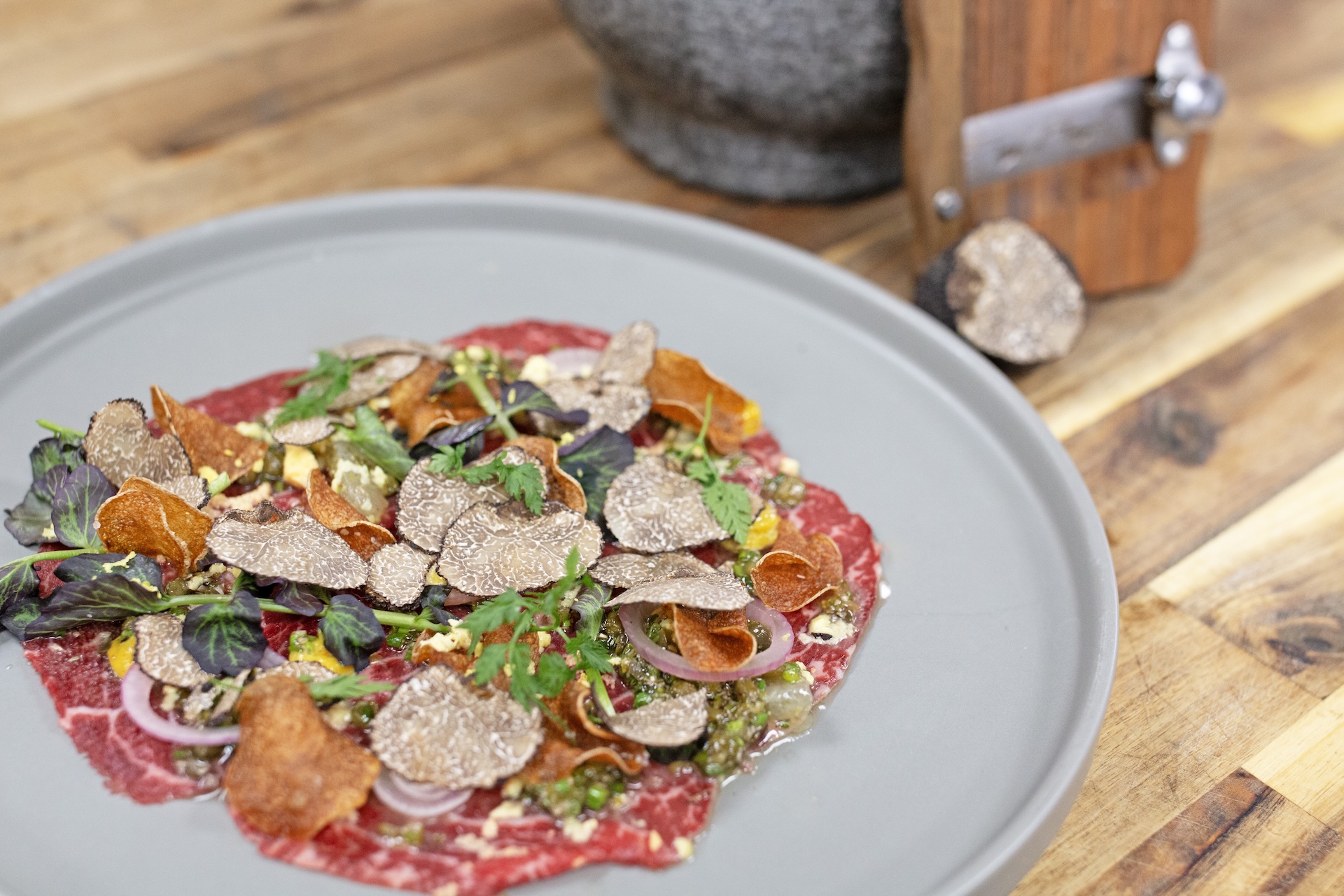 A gourmet dish of thinly sliced raw beef topped with shaved truffles, herbs, pickled onions, and crispy vegetable chips, served on a gray plate on a wooden table. A truffle and slicer are in the background.
