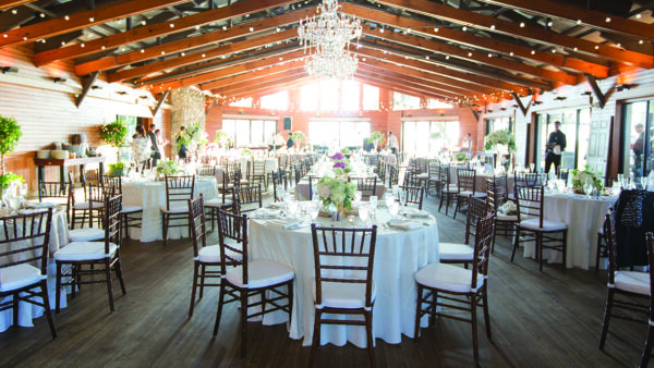 A spacious banquet hall set for an event, with round tables covered in white cloths, wooden chairs, flower centerpieces, string lights, chandeliers, and large windows letting in natural light.
