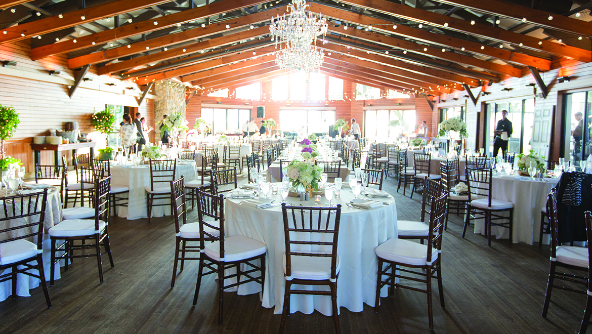 A spacious banquet hall set for an event, with round tables covered in white cloths, wooden chairs, flower centerpieces, string lights, chandeliers, and large windows letting in natural light.