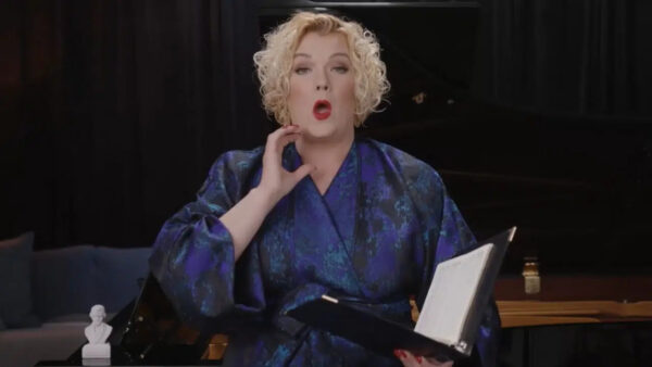 A person with curly blonde hair in a blue patterned robe appears to be singing, holding a black folder with sheet music. They are gesturing near their neck, with a piano and dark curtains in the background.