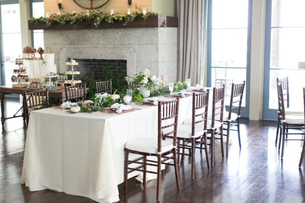 A bright, elegant dining area with a rectangular table covered in a white tablecloth, wooden chairs, and green and white floral centerpieces. In the background is a stone fireplace and dessert table. Natural light streams in from large windows.