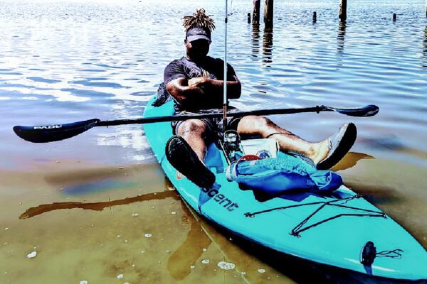 A man sits in a blue kayak on calm water, holding a paddle across his lap. He wears a cap and sunglasses, with a bag and gear in front of him. The shoreline and wooden posts are visible in the background.