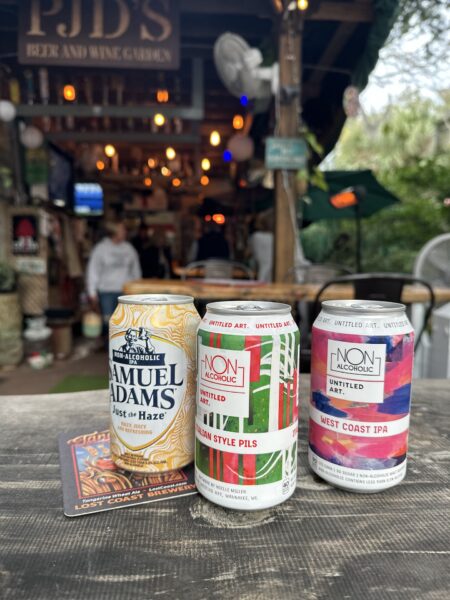 Three cans of non-alcoholic beer, including Samuel Adams Just the Haze and two Untitled Art varieties, sit on a table at an outdoor bar with string lights and greenery in the background.