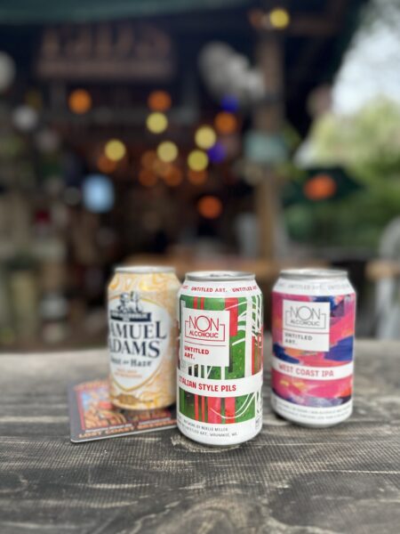 Three cans of craft beer, including Samuel Adams and two from Untitled Art (Italian Style Pils and West Coast IPA), are placed on a wooden table outdoors with a blurred, festive background.