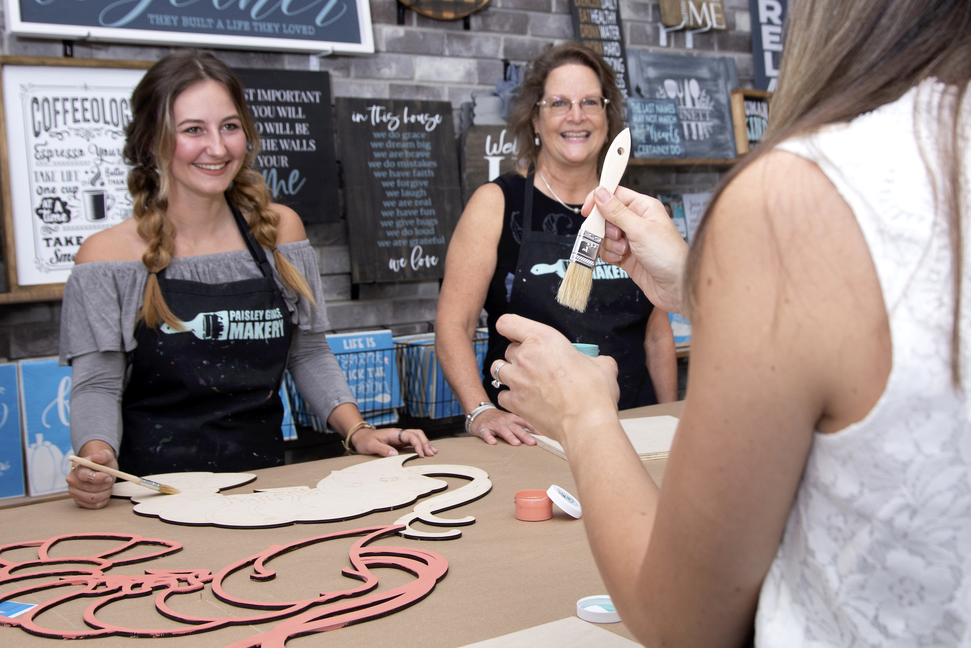Three women smiling and painting wooden crafts at a table in a workshop, with inspirational signs and art supplies displayed on the wall behind them.